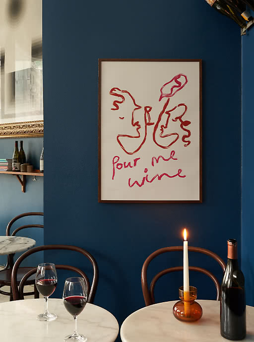 "Pour me wine" line-art poster in a wood frame on a deep blue wall above bistro seating — characterful wall art that sets the mood in restaurants and wine bars
