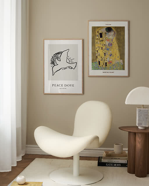 A Leo Gestel Peace Dove line drawing print and a Gustav Klimt The Kiss print displayed together above a sculptural white lounge chair, with a walnut side table and ribbed table lamp completing the corner.