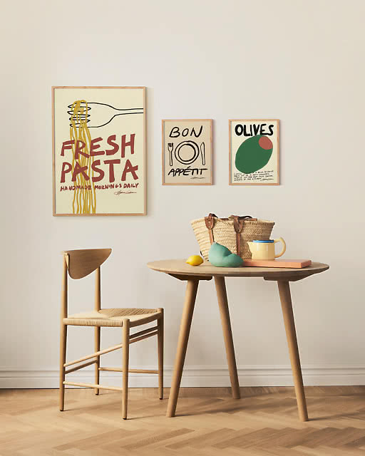 Three food-themed prints including Fresh Pasta, Bon Appétit, and Olives displayed together on a light wall above a round wooden dining table with a simple chair.
