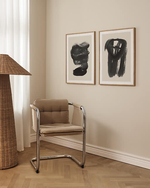 Two large abstract prints with expressive black brushstroke forms on warm beige backgrounds, displayed side by side in oak frames above a cantilever chair with chrome frame and a sculptural wicker floor lamp.