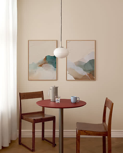 Two framed abstract prints in soft teal, blush and white tones hang above a round red café table with two wooden chairs, with a white ribbed pendant lamp centred between the prints.