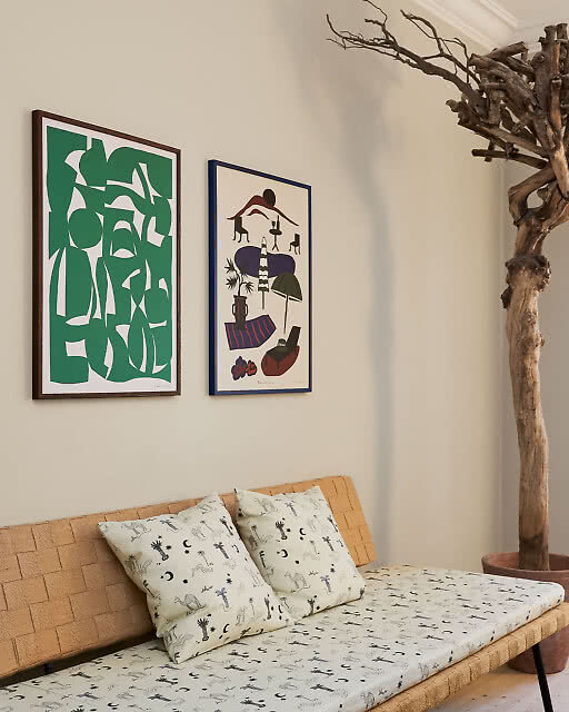 Two bold framed prints above a woven daybed — a vivid green abstract cutout print and a colourful illustrated still life — with a sculptural bare tree branch in a terracotta pot adding dramatic height to the corner.
