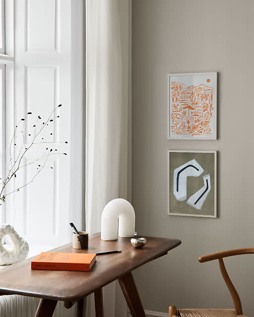Two framed prints in a styled desk corner — a detailed orange line-drawn Mediterranean landscape and a muted olive abstract geometric print — paired with a dark wood desk and a sculptural white arch lamp.