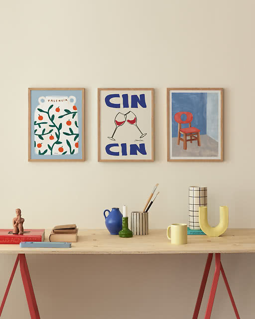 Three colourful framed prints — a playful pattern, a Cin Cin wine glass print, and a bold chair illustration — arranged on a white wall above a trestle desk with yellow and blue accessories.