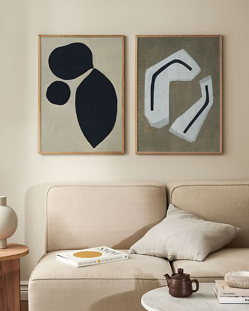 Two bold abstract prints displayed above a neutral linen sofa — one featuring dark organic shapes on a soft grey background and one with pale geometric forms on an olive green textured background.