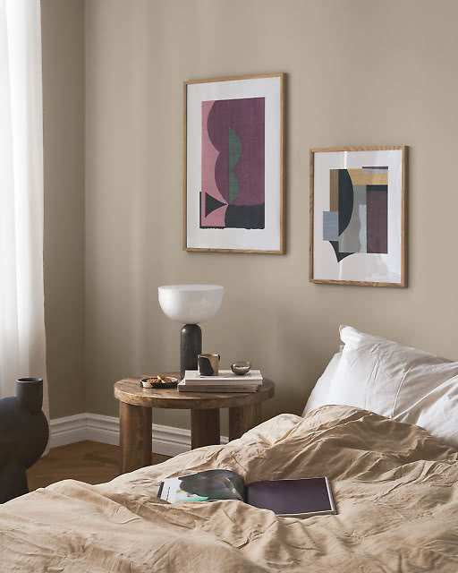 Two framed abstract prints in rich plum, dusty pink and deep green tones hang asymmetrically in a bedroom corner, paired with a sculptural black marble side table and a dome-shaped table lamp.