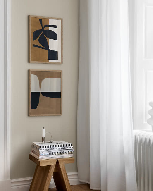 Two framed prints stacked vertically in a bright corner — a bold abstract botanical print in dark navy and amber tones and a graphic arch composition in navy and cream — beside a stool stacked with magazines and a candle.