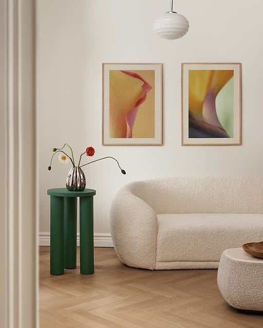 Two framed abstract prints in warm burgundy and olive tones displayed above a curved cream sofa, with a green side table and a single stem in a vase adding a pop of colour.