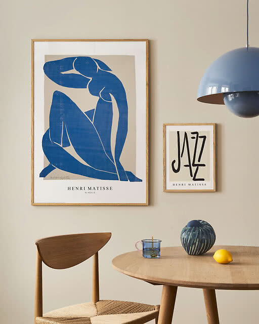 A large Henri Matisse Nu Bleu II print and a smaller Jazz print by the same artist displayed side by side in a Scandinavian dining room, above a round oak table with a blue dome pendant light.