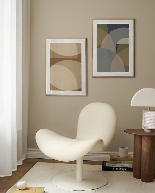Two framed abstract geometric prints in muted earth tones hung on a warm beige wall, paired with a sculptural white armchair and a wooden side table.