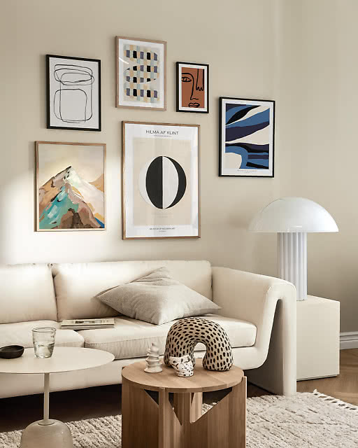 A curated gallery wall with five framed prints in warm neutral tones, styled above a cream sofa with a sculptural mushroom floor lamp and wooden side table.