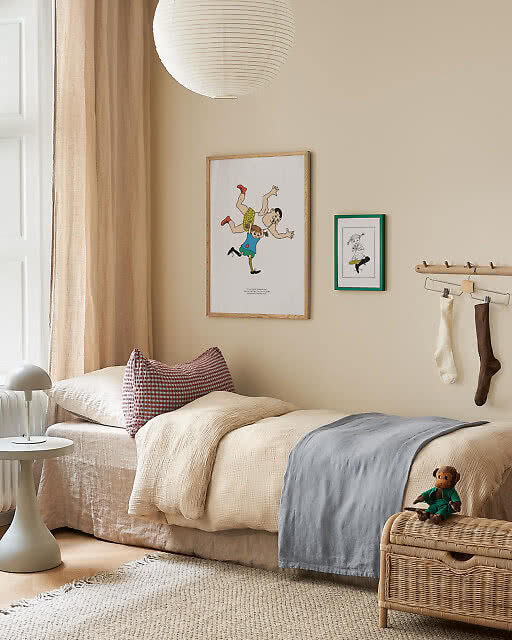 Two illustrated children's prints above a cosy bed — a lively tumbling figures scene in an oak frame and a smaller character illustration in a green frame — in a warm, softly lit child's bedroom.