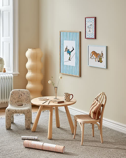 Three playful illustrated prints in a children's room corner, including a tightrope walker on blue stripes, a spotted zebra, and a tiger illustration, displayed above a small round play table with children's chairs.
