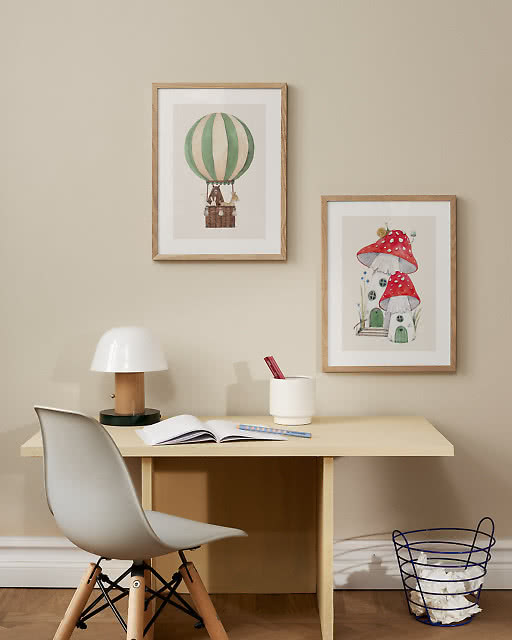 Two whimsical children's illustrations in oak frames above a pale yellow desk — a hot air balloon carrying animals and a red mushroom house — in a softly styled children's room.