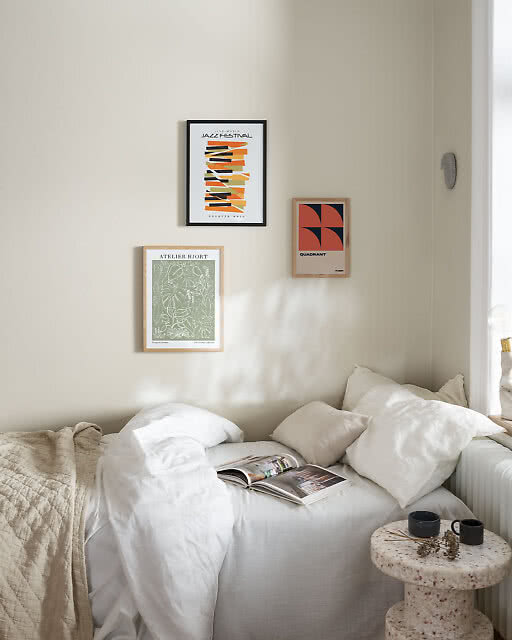 Three framed prints arranged in a corner of a softly lit bedroom — a Jazz Festival print with colourful abstract shapes, an Atelier Hjort botanical print in sage green, and a bold red and navy Quadrant geometric print — above a linen bed and terrazzo side table.