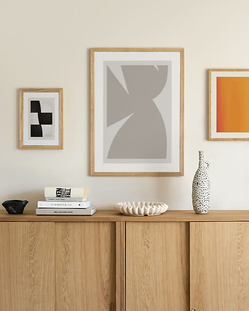 A trio of framed abstract prints in neutral and orange tones arranged above a light oak sideboard, accessorised with ceramic vases and decorative objects.