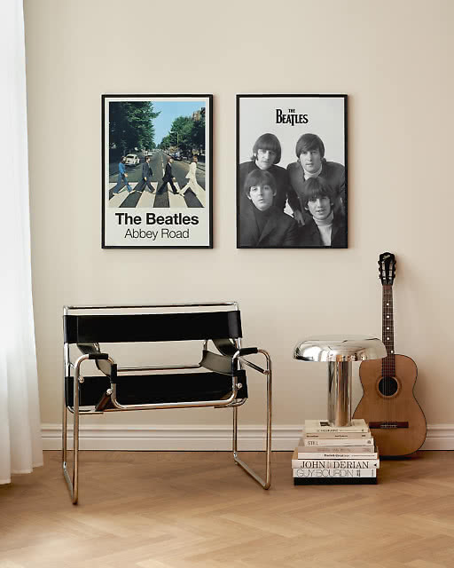 Two Beatles prints — Abbey Road and a band portrait — framed side by side on a white wall, styled with a Wassily chair, stacked books, and an acoustic guitar.