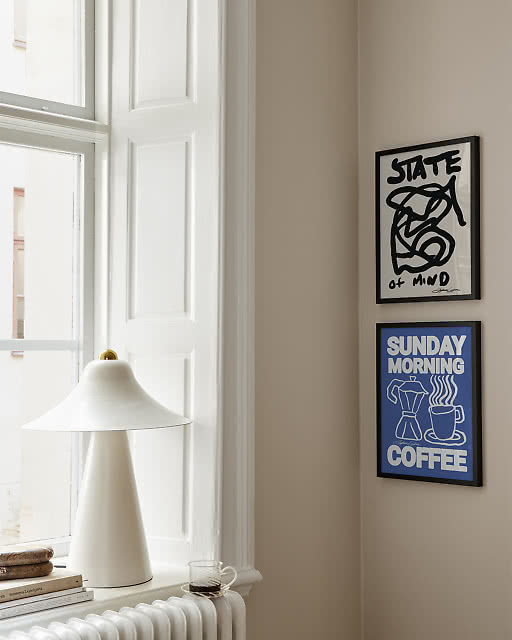 Two framed prints displayed on a warm beige wall next to a bright window — a bold black abstract line art print reading State of Mind and a vibrant blue typographic print reading Sunday Morning Coffee, styled with a white conical table lamp.