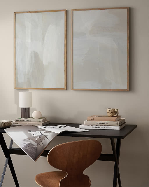 Two soft abstract prints in pale grey and white tones hung above a dark desk, styled with a table lamp, stacked books, and a warm wooden chair.