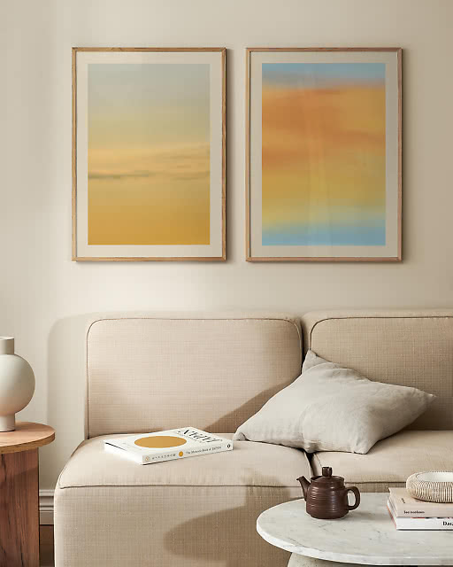 Two framed prints displayed on a warm beige wall next to a bright window — a bold black abstract line art print reading State of Mind and a vibrant blue typographic print reading Sunday Morning Coffee, styled with a white conical table lamp.