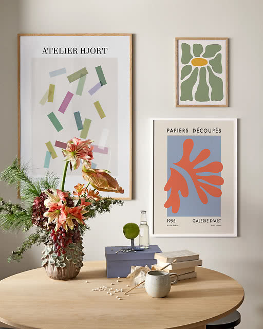 Three prints above a round table with a dramatic floral arrangement — a confetti Atelier Hjort print, a green daisy, and a Papiers Découpés red coral cutout on blue.