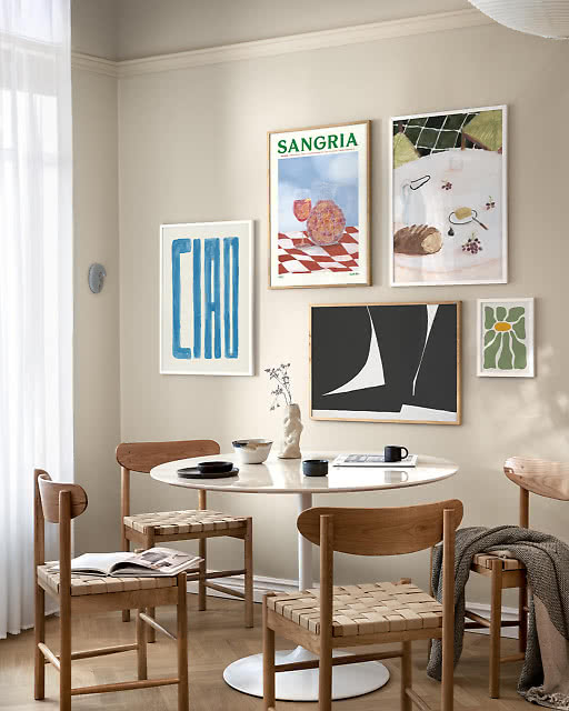 A five-print gallery wall in a Scandinavian dining room, featuring a Ciao typographic print, a Sangria illustration, a still life with bread and fruit, a bold black and white abstract, and a small green daisy print — all arranged above a white tulip table with woven oak chairs.