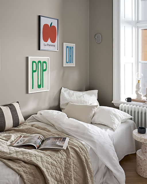 Three typographic and fruit prints displayed in a bedroom corner — a bold green Pop print, a La Pomme apple illustration, and a blue Ciao print — above a linen bed with white pillows and a quilted throw.