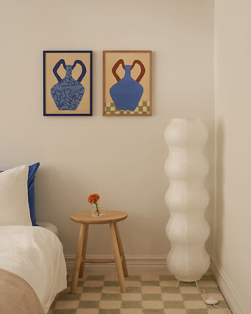 Two illustrated amphora vase prints in warm sandy tones — one with a textured blue pattern in a navy frame, and one with a smooth blue body and terracotta handles on a checkerboard base in an oak frame — displayed beside a sculptural bubble floor lamp in a bedroom.