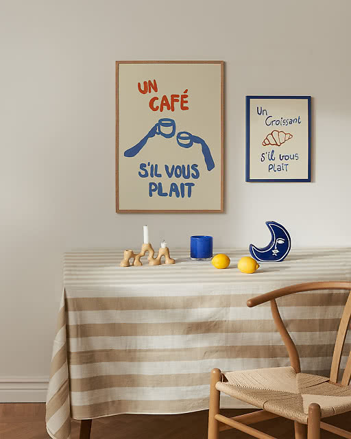 Two French café-themed prints displayed above a striped linen tablecloth — a bold Un Café S'il Vous Plaît illustration in red and blue, and a smaller Un Croissant S'il Vous Plaît print in a navy frame — styled with candleholders, lemons and a crescent moon ceramic.