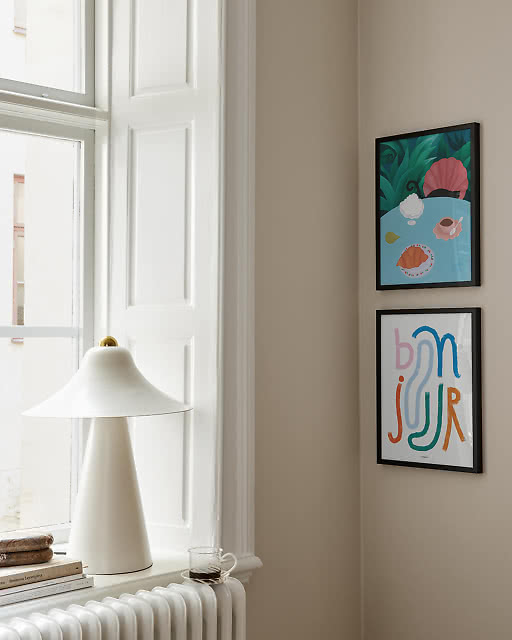 A colourful tropical still life print and a multicoloured Bonjour typographic print in black frames beside a bright window.