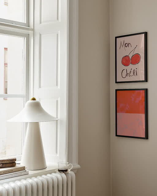 A playful Mon Chéri cherry illustration print and a bold red-orange colour block print displayed in black frames beside a bright window, styled with a white conical table lamp on the windowsill.