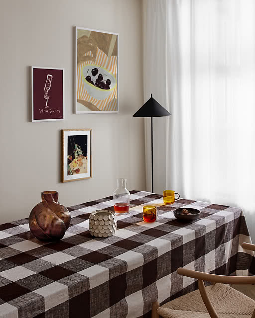 Three prints displayed above a dark check tablecloth dining table — a Vino Pouring illustration in burgundy, a colourful cherry bowl still life, and a small night market scene — alongside a black conical floor lamp and amber glassware.