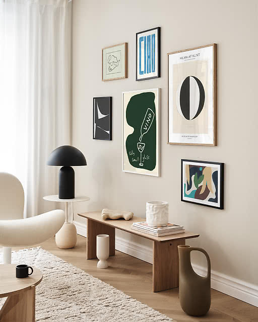 A seven-print gallery wall in a light Scandinavian living room, including a Ciao typographic print, a Hilma af Klint poster, a dark Vino bottle illustration, a small abstract, a line drawing, and a colourful figurative print, styled above a wooden bench with ceramic vessels.