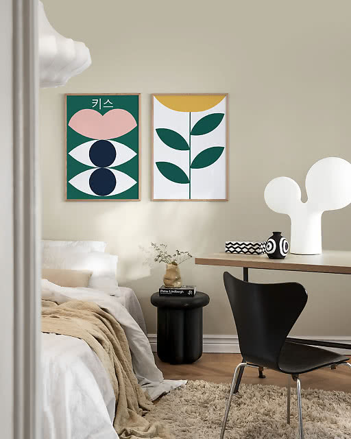 Two graphic prints above a bed — a bold green print with Korean text and abstract eye and lip motifs, and a minimal botanical stem with green leaves and a yellow sun — in a calm bedroom with a black Arne Jacobsen chair and sculptural white lamp.