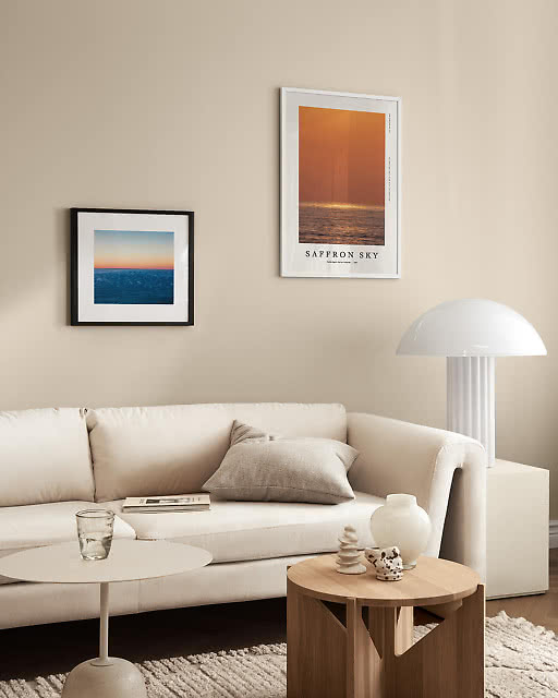 A Saffron Sky ocean sunset print in a white frame and a smaller blue seascape photograph in a black frame displayed above a curved cream sofa, with a white dome lam