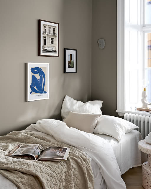 Three framed prints arranged in a moody sage green bedroom corner — a Henri Matisse Nu Bleu print, a black-framed architectural photograph of a white Porsche outside a townhouse, and a small colourful interior photograph — above a linen bed with an open magazine.