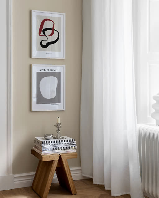 A bold red and black abstract line print and an Atelier Hjort grey oval print, stacked above a stool with magazines.