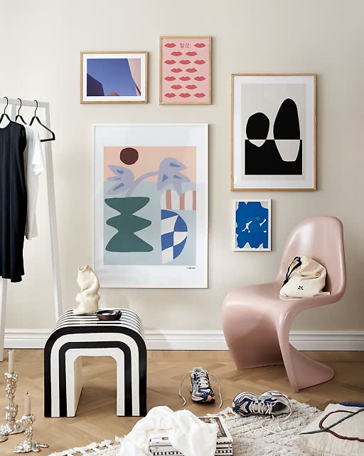 Five-print gallery wall with a large pastel abstract as centrepiece, flanked by a lips print, blue photo, and abstract shapes, with a pink Panton chair.