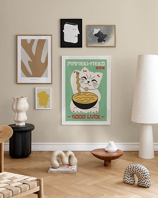 five-print gallery wall featuring a bold Maneki-Neko Good Luck cat ramen illustration as the centrepiece, surrounded by a tan leaf cutout print, a small yellow botanical, a quirky illustrated face, and a grey snow scene, in a warm living room with sculptural objects.