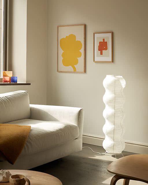 A bold yellow flower print and a small red geometric abstract displayed above a cream sofa, beside a tall sculptural white paper floor lamp.