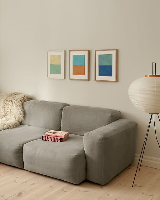 Three small framed colour-block prints in yellow-green, orange-teal and blue-green tones hang in a row above a deep grey modular sofa, with a paper lantern floor lamp adding warmth to the side.
