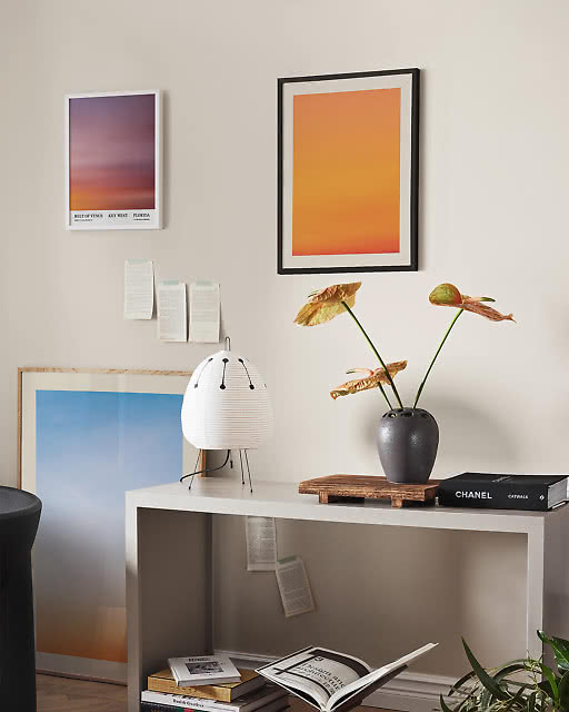 A styled workspace corner featuring a Belt of Venus Key West sunset print, a large vibrant orange gradient print in a black frame, and a soft blue sky print leaning against the wall, alongside a paper lantern table lamp and anthurium in a ceramic vase.
