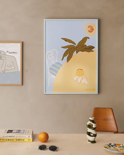 A large light blue-framed illustrated print by Isabelle Feliu showing a sunny scene with a palm plant, peeled fruit and a striped ball.
