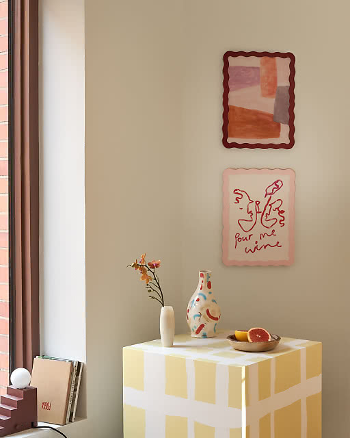 Two framed prints with wavy red and pink borders displayed above a yellow check sideboard — a soft abstract colour block print in terracotta and mauve tones, and a playful Pour Me Wine line drawing — with ceramic vases and a halved grapefruit styled below.