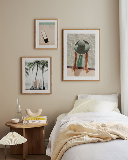 Three summer photo prints — a pool scene, surfboards under palms, and a collage — above a bed with a wooden side table.