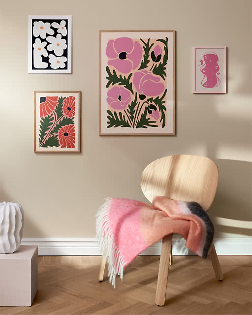 Four botanical prints in pink and red tones — large pink poppies as centrepiece — above a wooden chair with a pink throw.