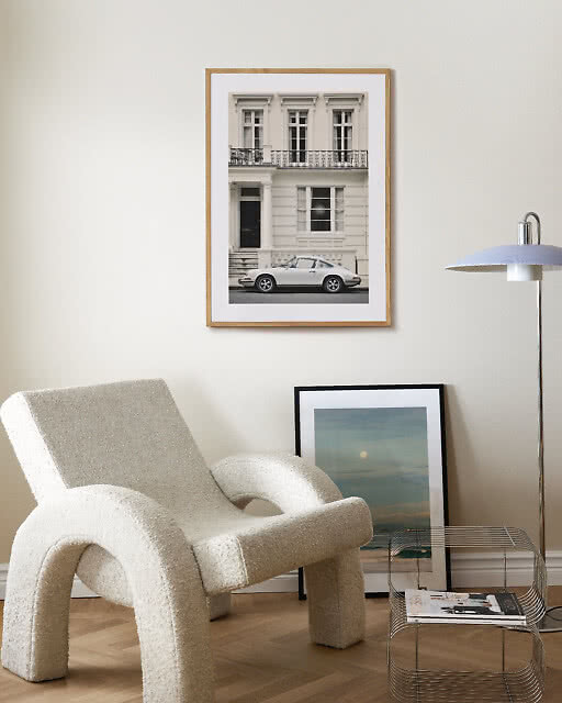 A black-and-white photographic print of a classic white Porsche parked in front of an elegant townhouse façade, hung above a sculptural bouclé armchair with a teal ocean print leaning against the wall below.
