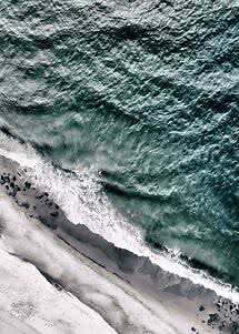 Aerial view of waves crashing onto a dark sandy beach with black rocks