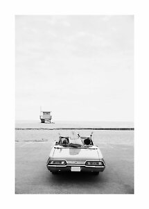 Black-and-white photo of two people on a car with a surfboard by the beach and a lifeguard tower beyond