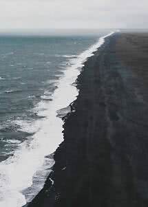 Luftfoto af bølger langs en lang sort sandstrand under overskyet himmel